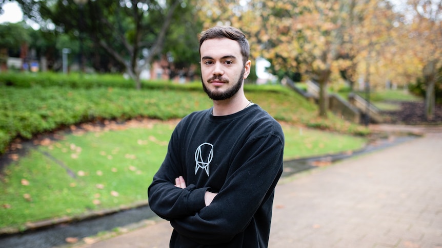 Young white man with dark short hair and facial hair faces the camera with a neutral expression in a park. his arms are crossed 