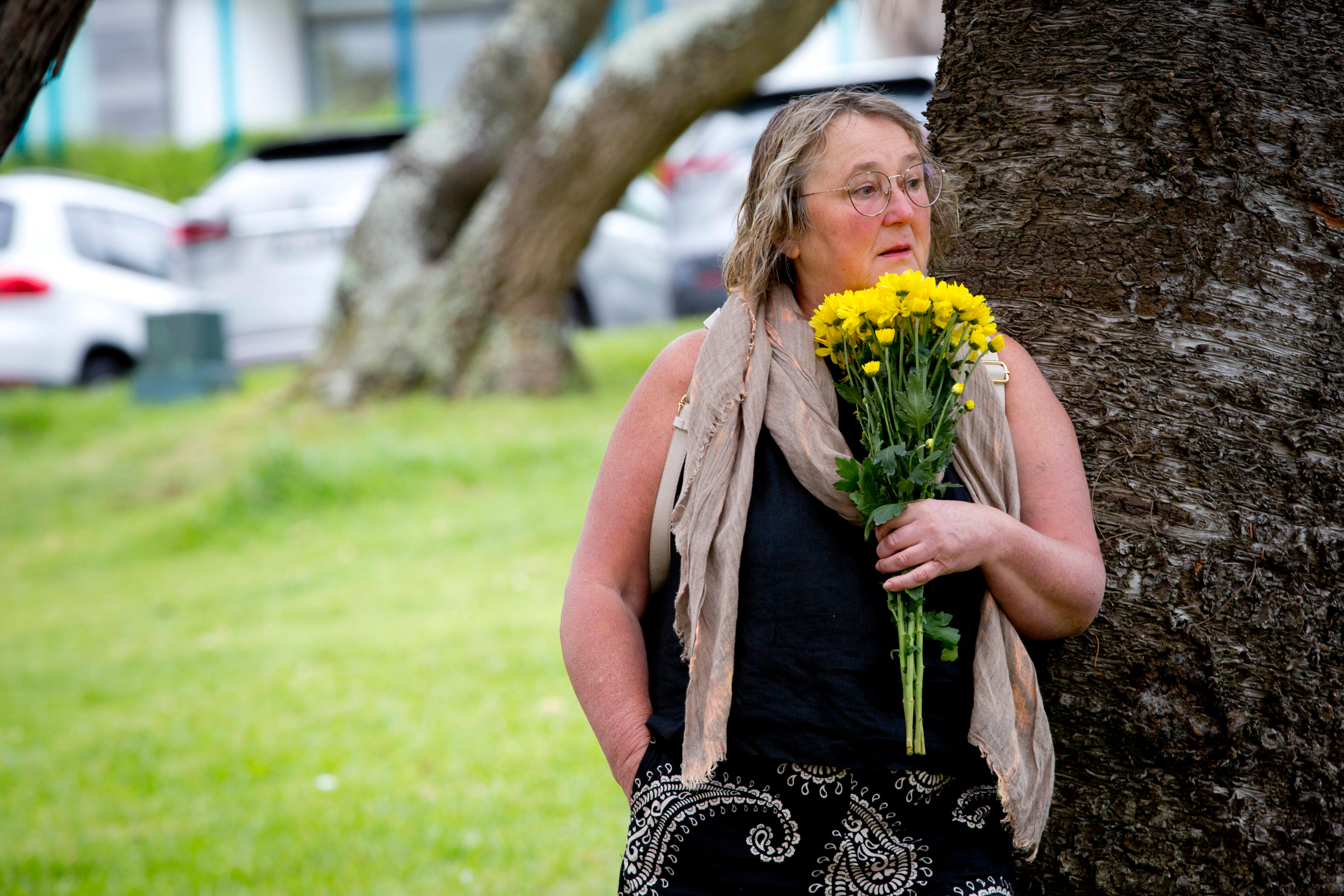 Uma mulher segura um ramo de flores amarelas e se encosta em uma árvore, olhando além da câmera
