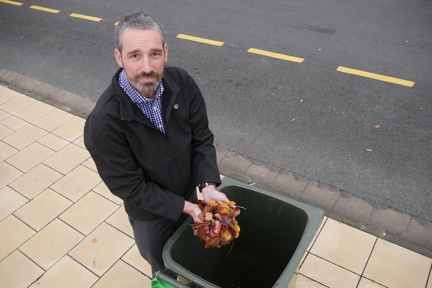 Man looking at camera with leaves in hands above organics bin on footpath.