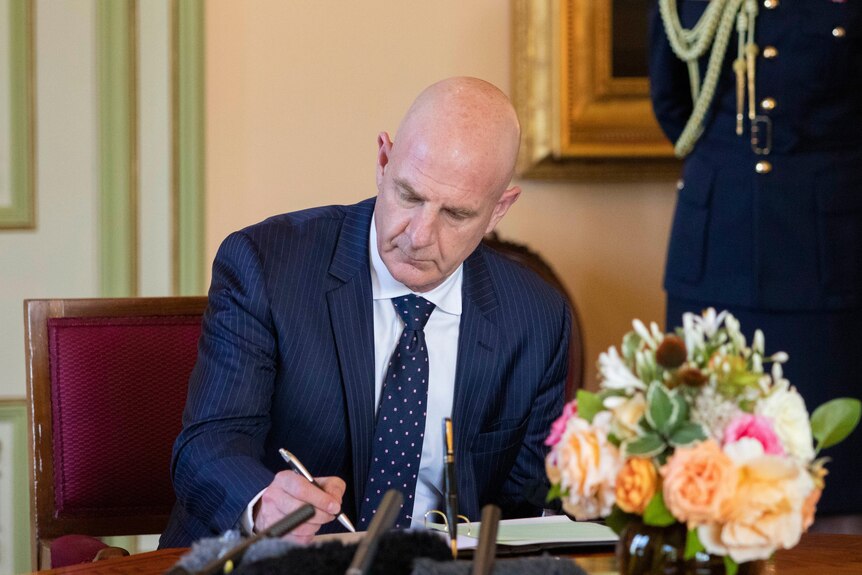 Bald man writes in a book sitting at a desk. 