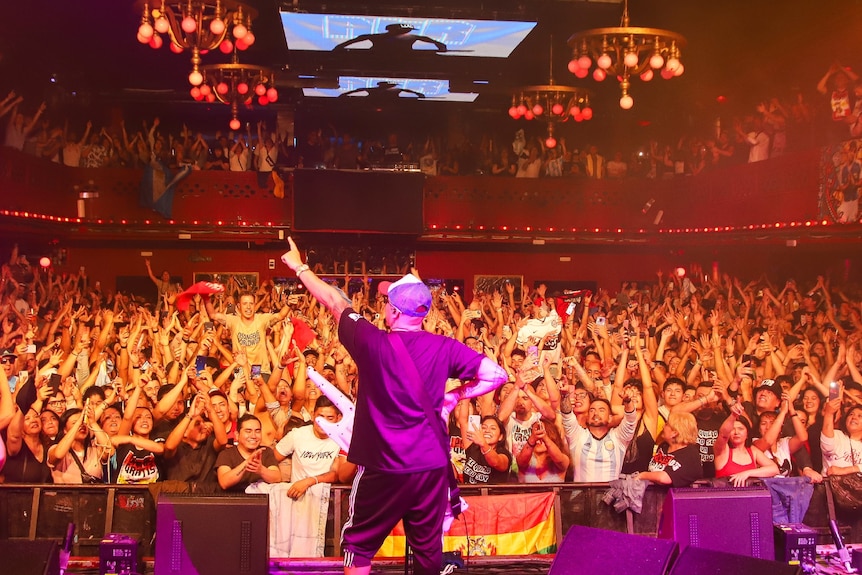 Man on a stage with arms in the air, with hundreds in the crowd, and people on the balconies.