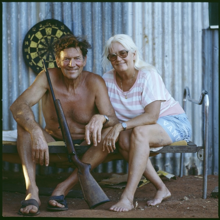 A couple sit outside a tin shed and smile at the camera, a shot gun leaning on the man.