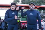 Two Indigenous men in westuits stand next to a SCUBA dive boat smiling