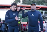 Two Indigenous men in westuits stand next to a SCUBA dive boat smiling