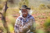 Indigenous elder wearing a black hat and checkered shirt, sitting on country.