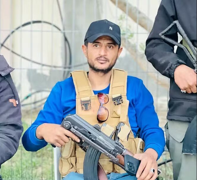 A young Palestinian man in a shirt, military vest and baseball cap holds what appears to be an automatic rifle.
