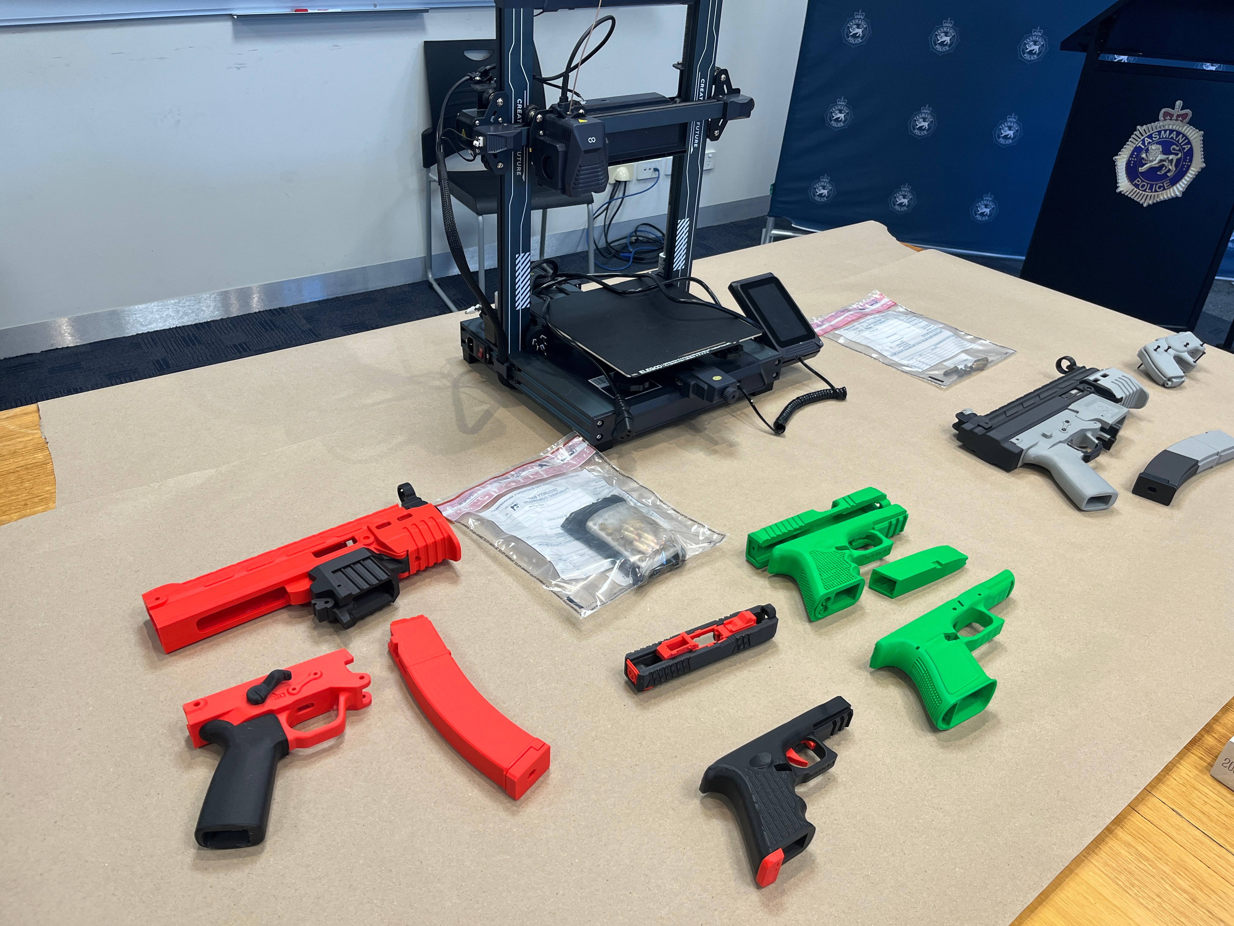 A dozen 3D printed firearms and a 3D printer laid out on a wooden table.