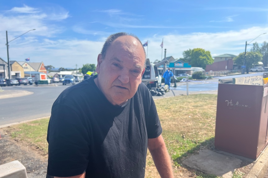 A middle-aged man with a sad look on his face stands in a country town beneath a sunny sky.