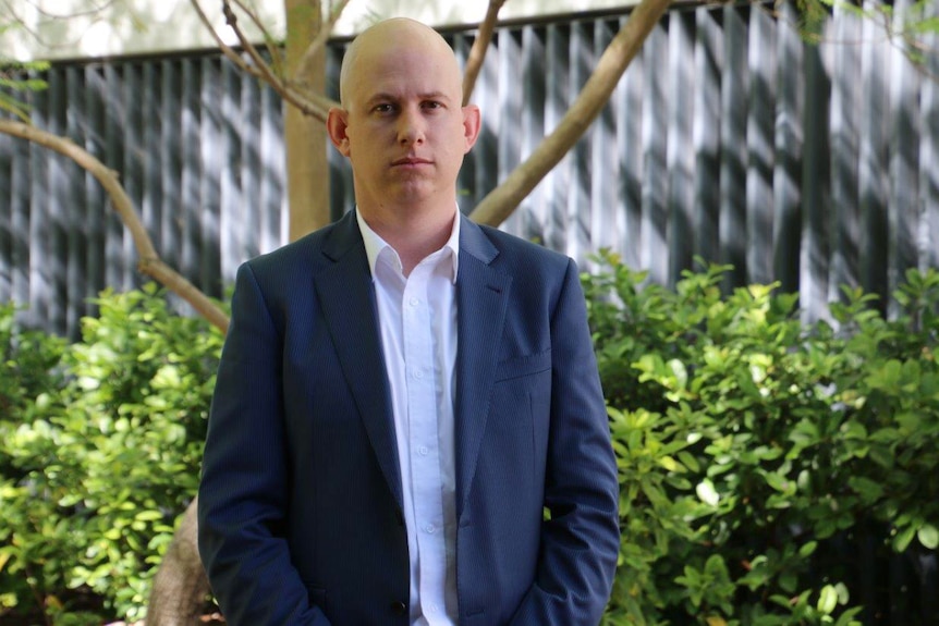 A mid-shot of SDA WA assistant secretary Ben Harris standing in front of a garden wearing a blue suit and white shirt.