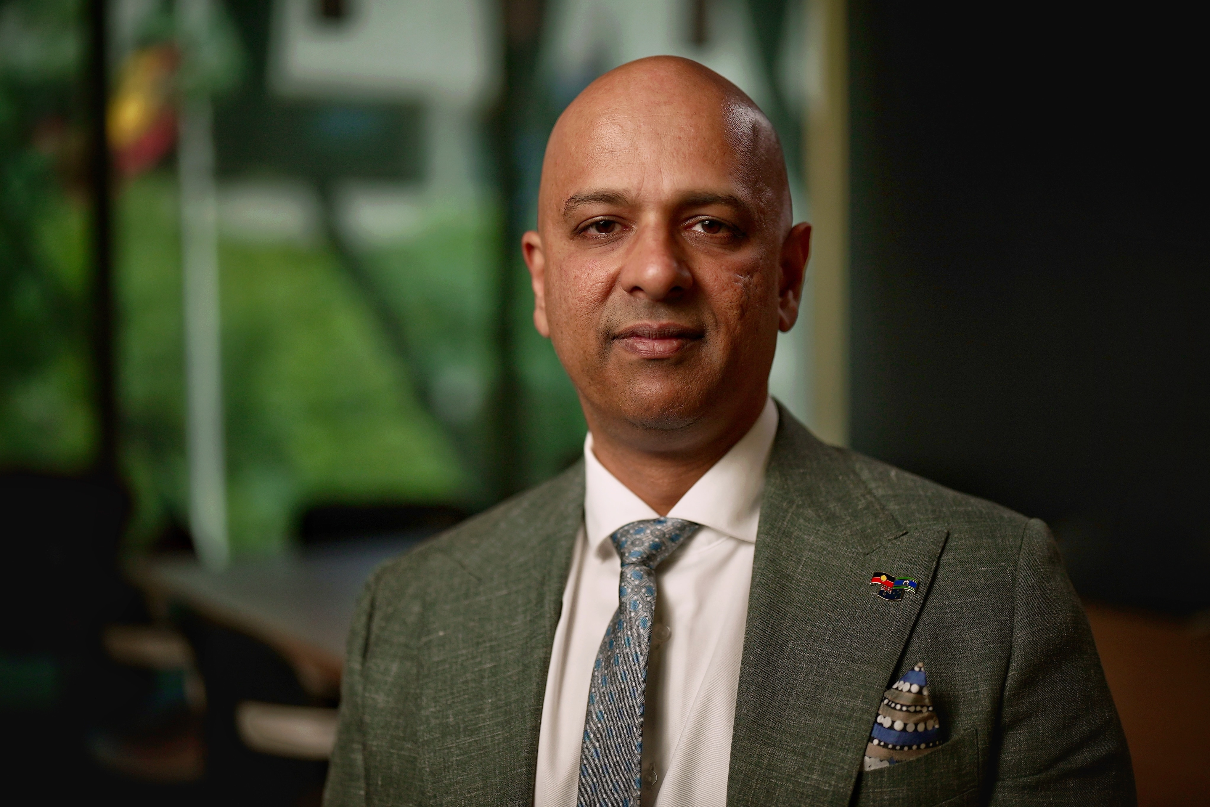 A man with a bald head wearing a suit and tie stands in front of a large meeting table and looks neutrally at the camera.