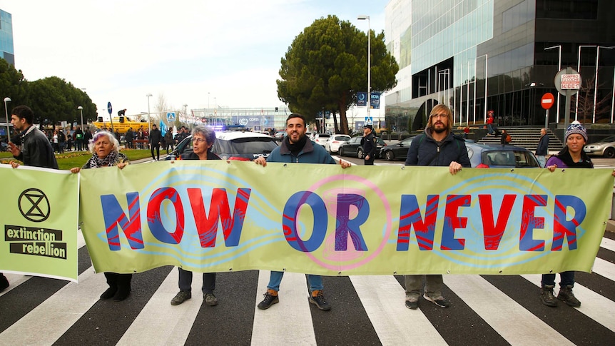 People hold a sign in the street that reads 'Now or Never'.