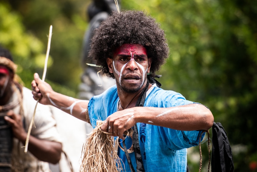 A Polynesian man with Afro hair and blue shirt with painted face holds stick and straw in dance pose