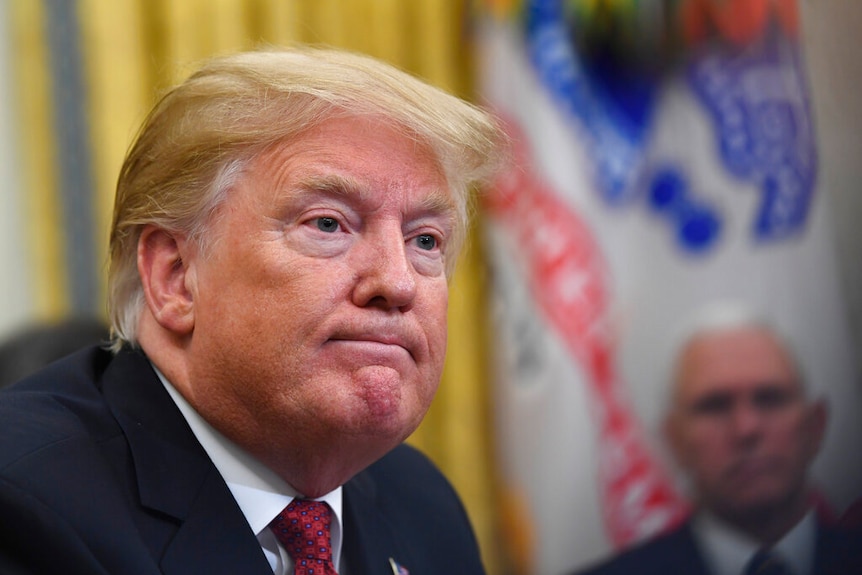 Donald Trump stares out of the frame with a mild grimace. Mike Pence can be seen in the blurred background.