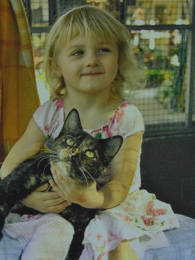 Blonde haired little girl with blue eyes nursing tortoiseshell cat.