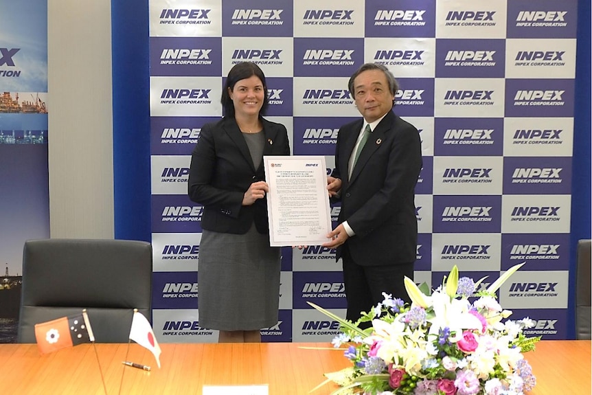 A smiling woman and man hold a signed document in a boardroom, with INPEX logo behind