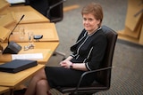 A fair-haired woman in a black suit with white piping detail, sits in an office chair in Scotland's parliament.