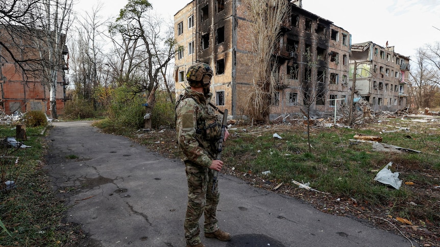 A man stands in camouflaged gear looking at a burnt out building.
