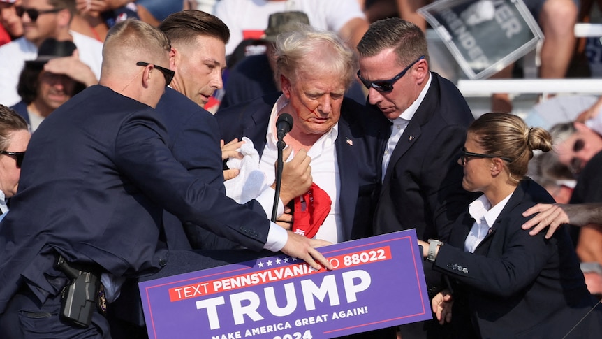 Trump surrounded by suited men and a woman, his shirt is open and you can see a little blood on his face