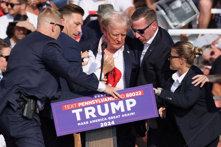 Trump surrounded by suited men and a woman, his shirt is open and you can see a little blood on his face