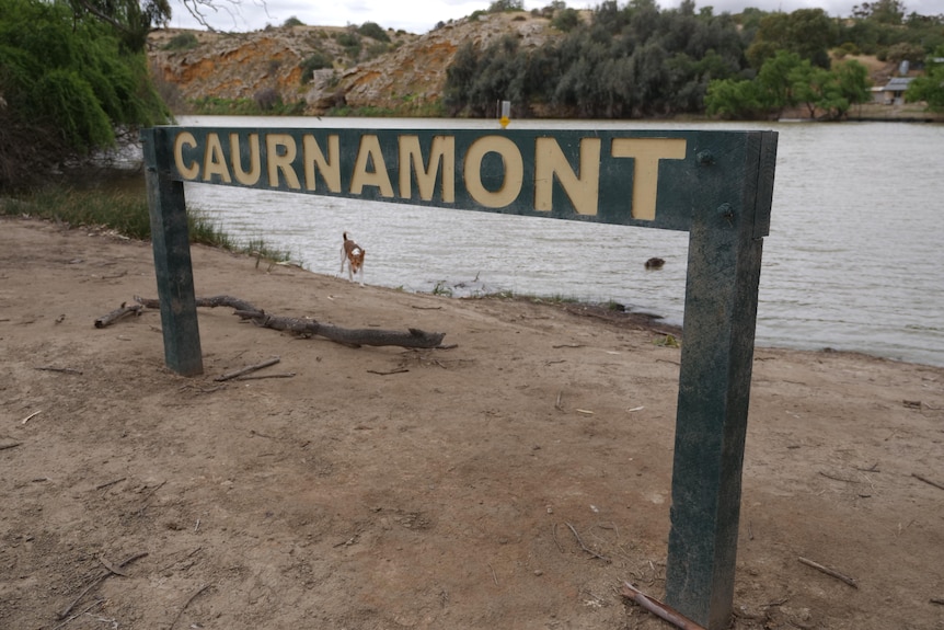 Green wooden sign, yellow writing reads CAURNAMONT. Sits on River banks with dark river behind it