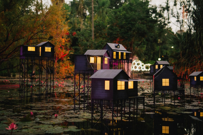 Une série de modèles de maisons Queenslander en aluminium dominent un lac avec une lumière chaude émanant de chacune d'entre elles.