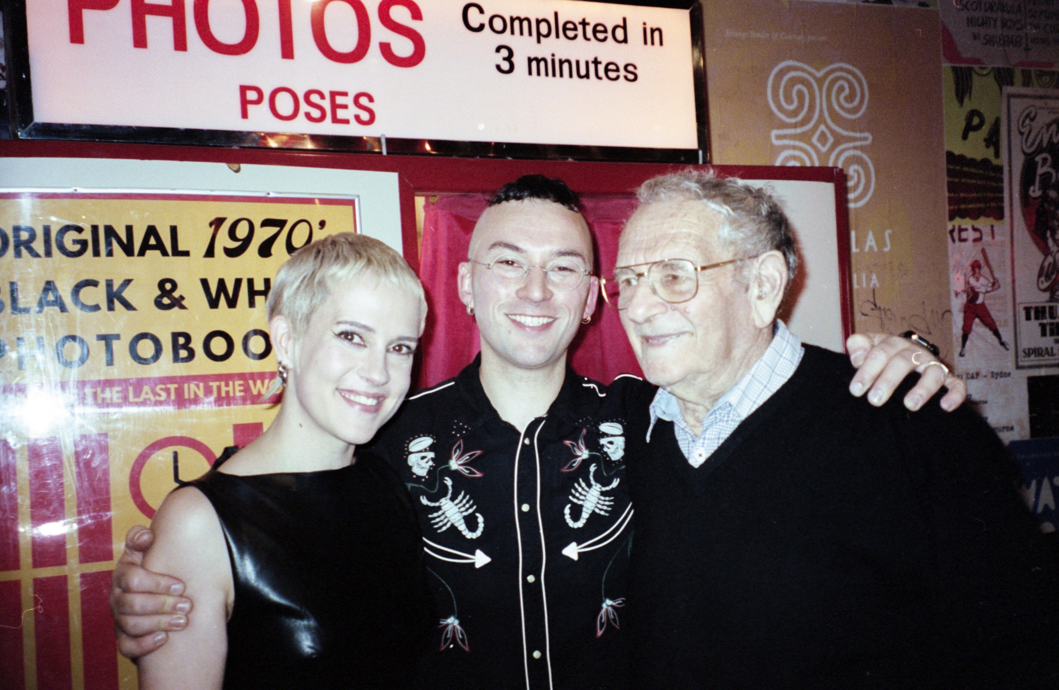 A young man and woman pose with their arms around an older man in front of a retro photo booth in a pub.