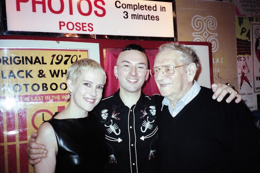A young man and woman pose with their arms around an older man in front of a retro photo booth in a pub.