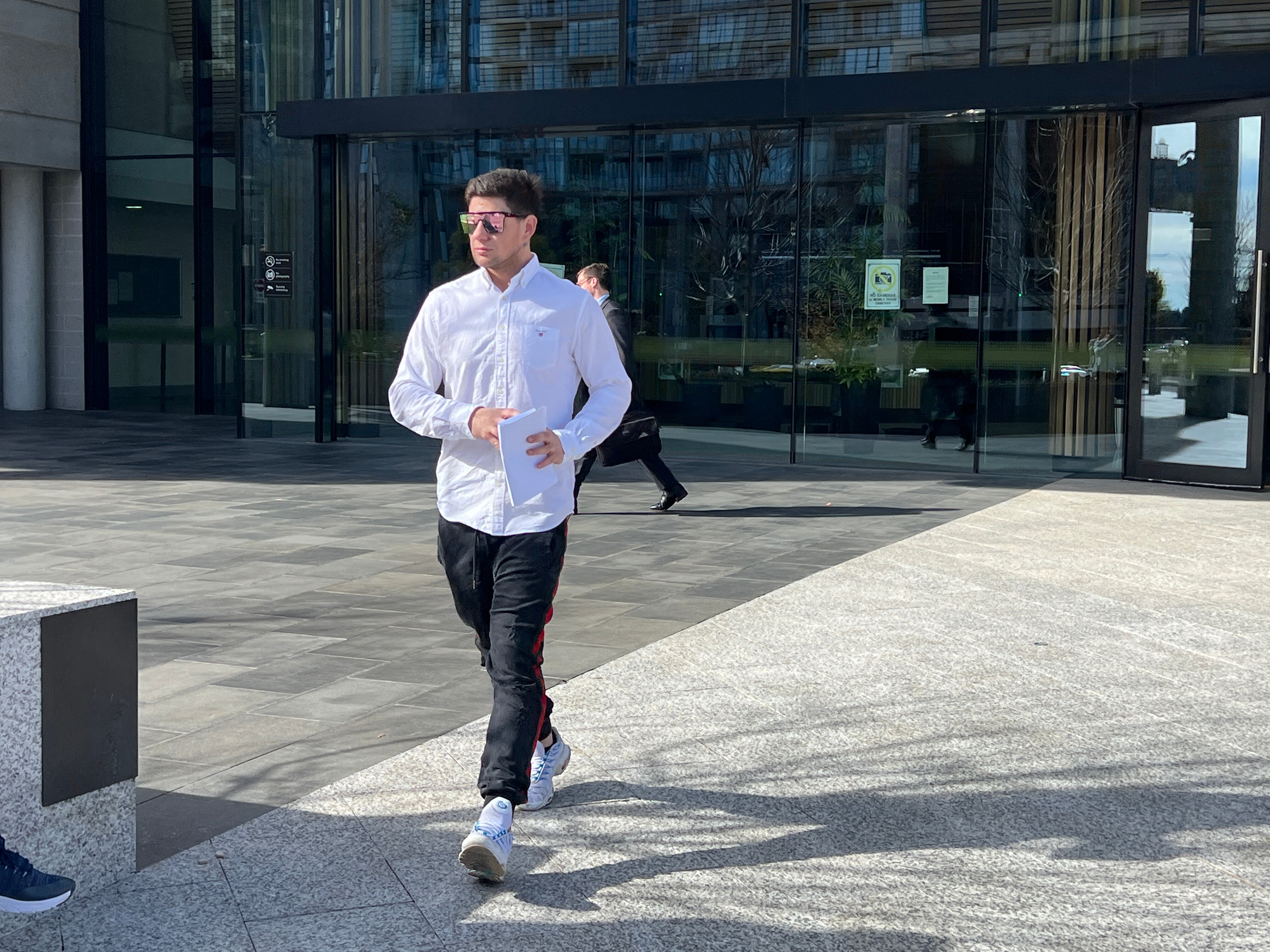 A man wearing a white button down shirt, black jeans and large sunglasses walks out of a court building holding papers.