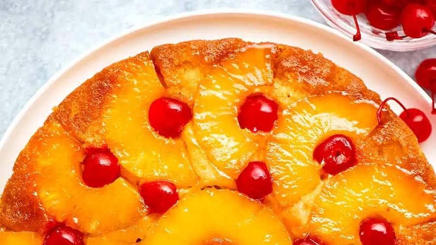 Bird's eye view of bright yellow and red pineapple cake, dotted with red cherries, on a white plate.