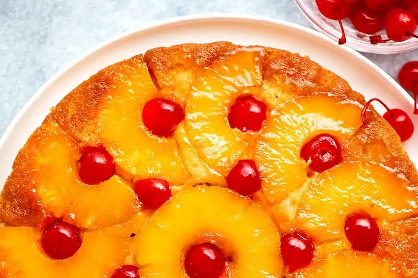 Bird's eye view of bright yellow and red pineapple cake, dotted with red cherries, on a white plate.
