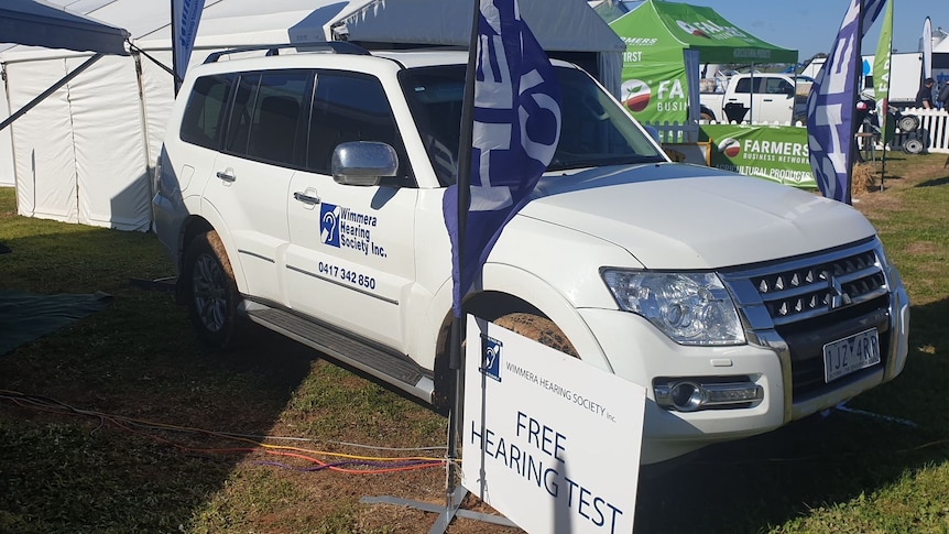 A white four wheel drive with blue insignia stands behind two flags while Field Day marquees stand in background 