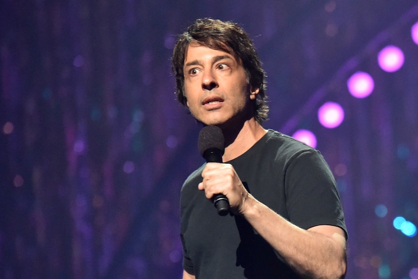 Arj Barker speaks into a microphone while standing on stage in front of purple backdrop