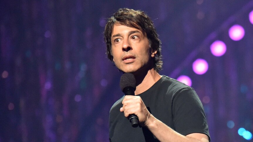 Arj Barker speaks into a microphone while standing on stage in front of purple backdrop