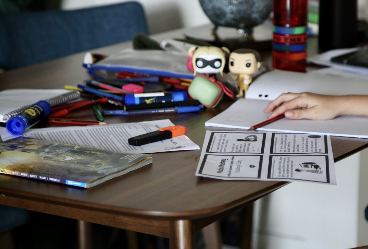 A child's hand holds a pencil next to school books and toys.