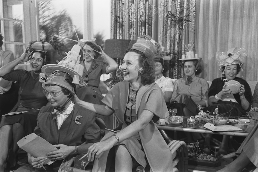 A black and white shot of laughing women wearing hats made out of Tupperware.