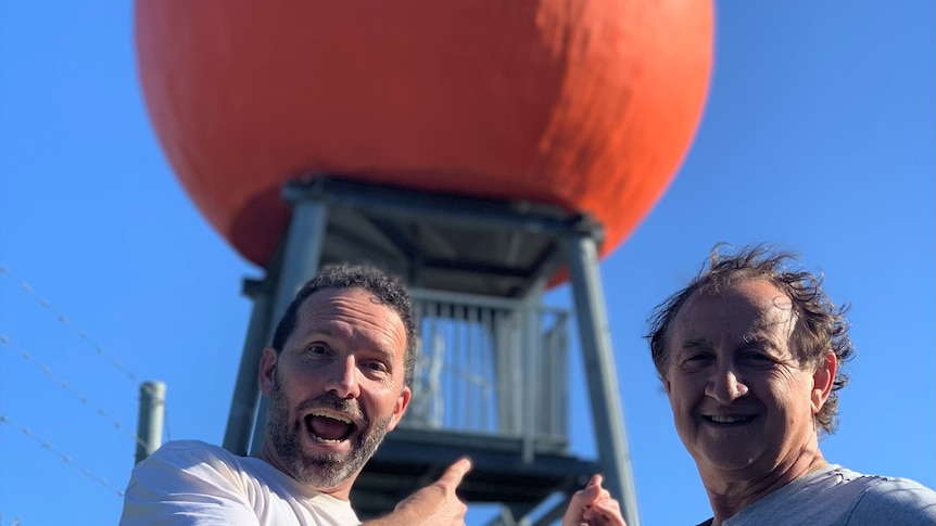 Excited man points at large orange statue as man smiles next to him