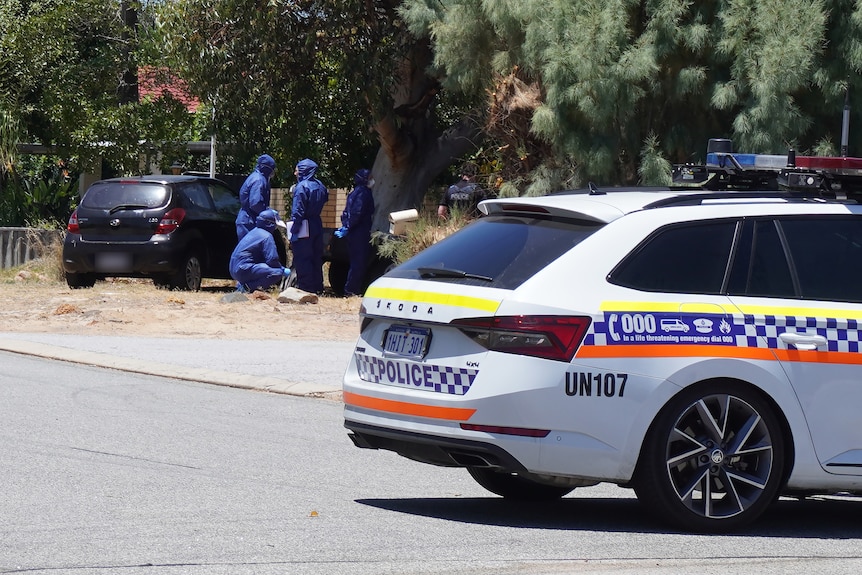 Police forensic officers in blue suits stand in the front yard of a house with a police car on the road in the foreground.