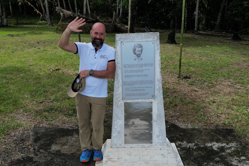 A man in a white polo shirt, Nicholay Miklouho-Maclay Jr, stands next to a monument and waves.