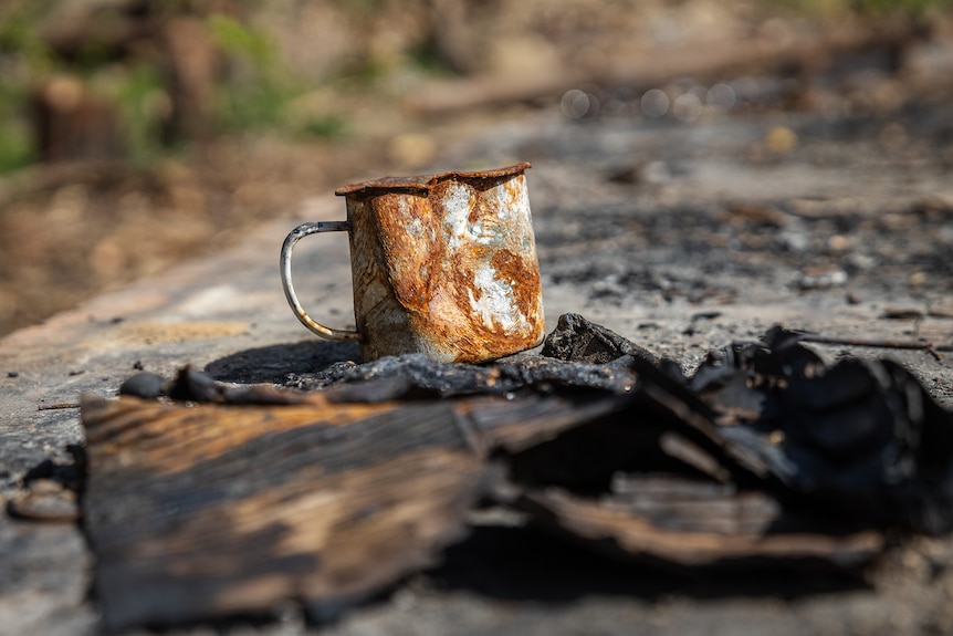 A metal mug scarred by fire sits on a bed of ash