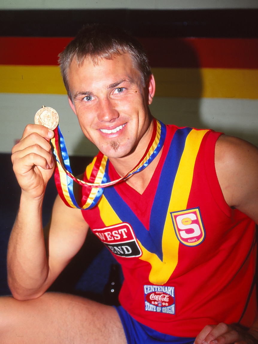 A smiling Australian rules player wearing a South Australian State of Origin guernsey holds up a medal f9or best on ground.