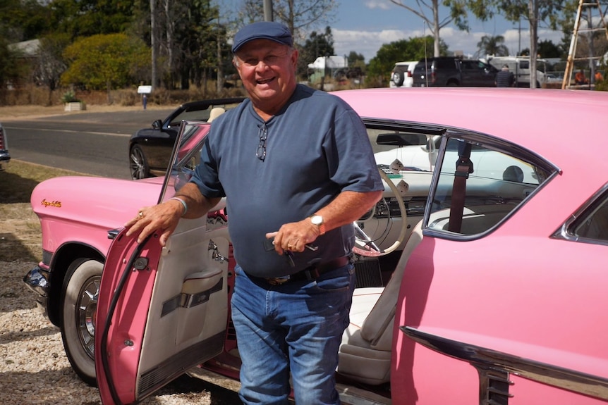 A man with a hat on, getting out of a pink vintage car, holding the door open and smiling