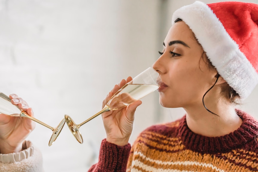 A young woman drinking a glass of champagne while wearing a santa hat.