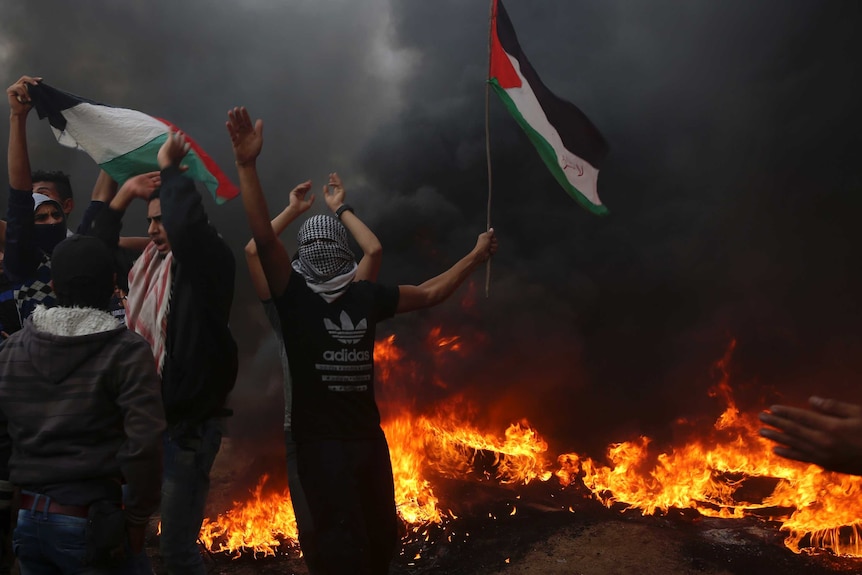 A man with a scarf on his face lifts his hand and holds a Palestinian flag among fire and smoke in Gaza.