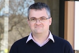 A man looks at the camera and stands in front of three news microphones. A council sign is in the background.