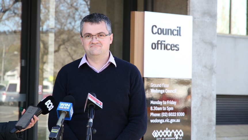 A man looks at the camera and stands in front of three news microphones. A council sign is in the background.