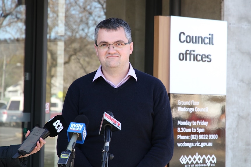 A man looks at the camera and stands in front of three news microphones. A council sign is in the background. 