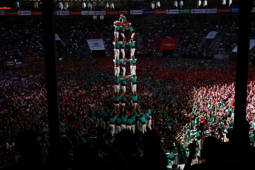 A group of people in teal shirts form a large human tower in the middle of a packed arena.