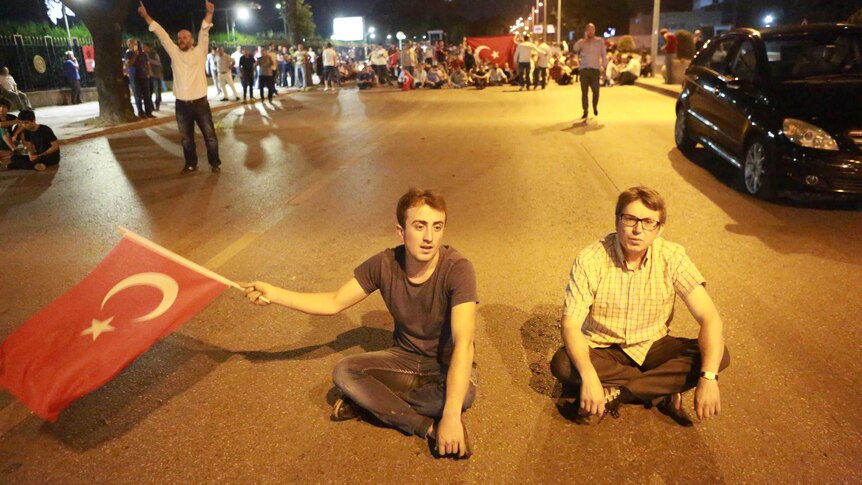Two men sit in the street, one of them waving a Turkish flag. A large crowd of protesters are in the background.