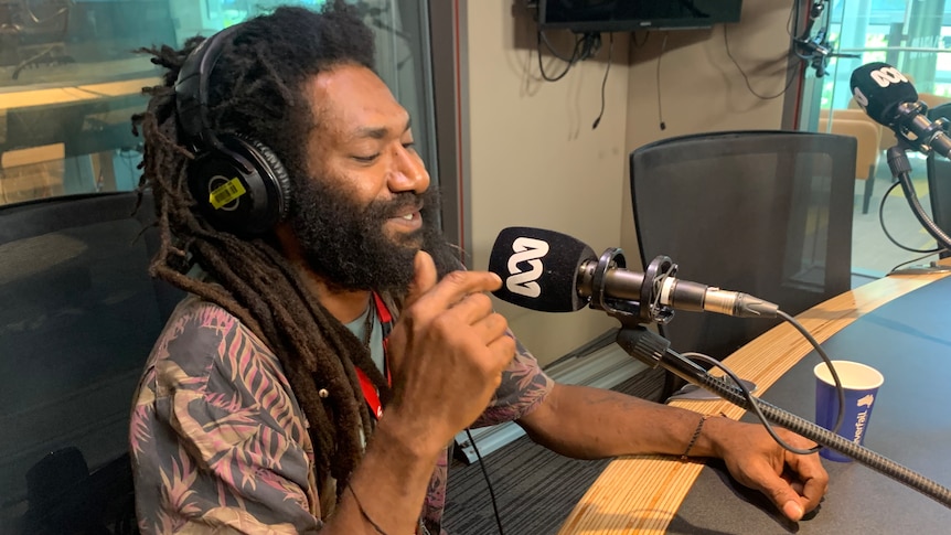 A man sits and smiles in front a microphone to the right of screen 