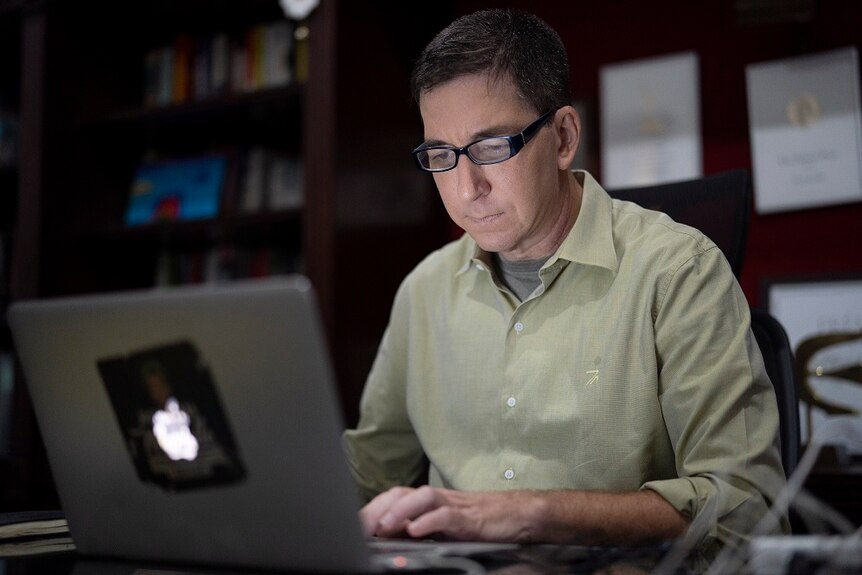 Journalist Glenn Greenwald sitting on a desk with a laptop.